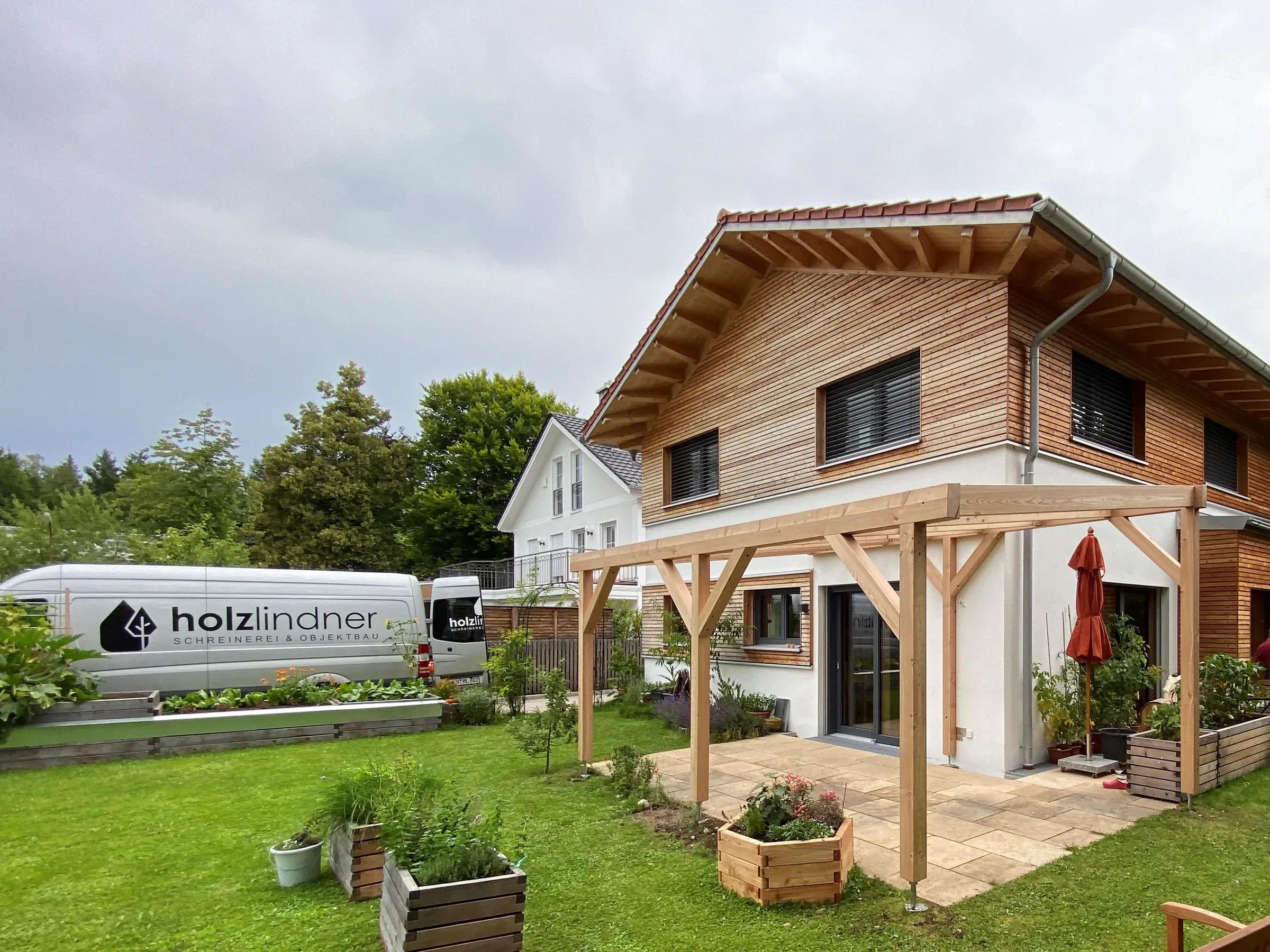 Wohnhaus mit waagerechter Holzschalung im Obergeschoss, Holzpergola über der Terrasse und Hochbeeten