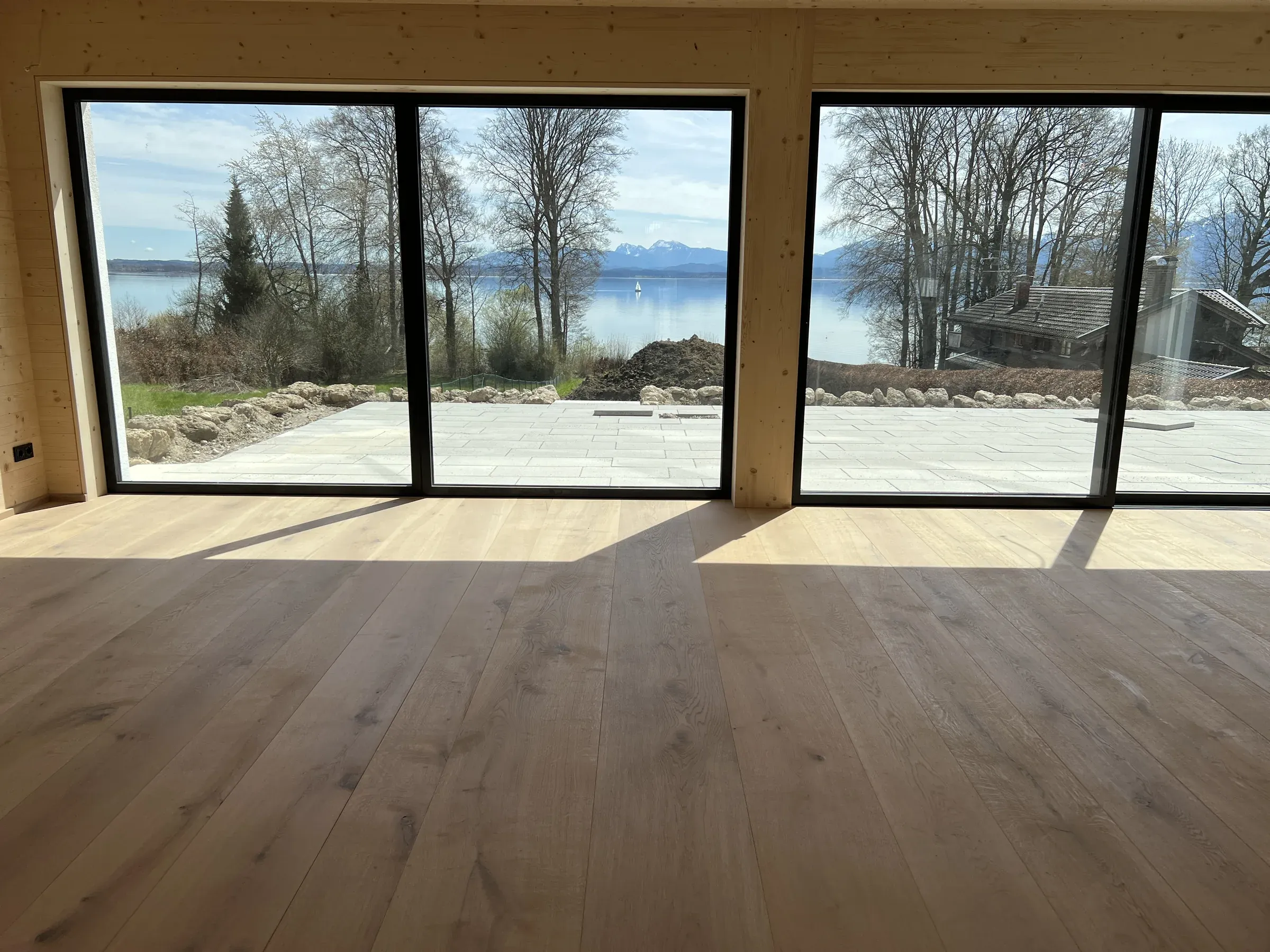 Wohnraum mit breiten hellen Landhausdielen, Holzwänden und großen dunkel gerahmten Fenstertüren mit Blick auf einen See und Berge